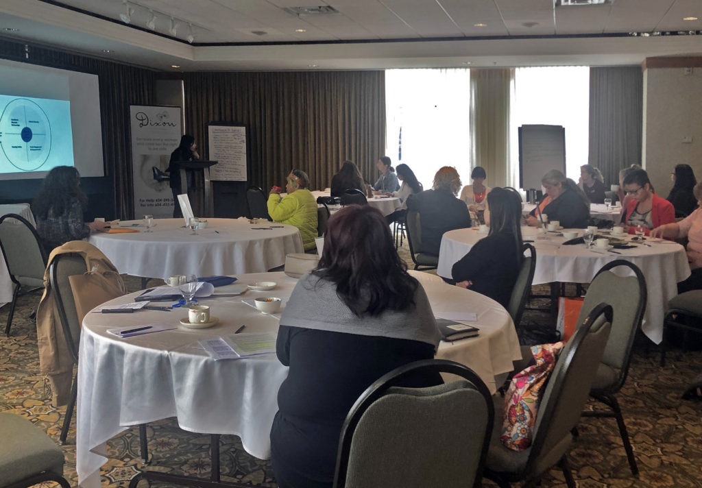 Transitional housing service providers seated around multiple round tables while Dixon's Housing Outreach Worker, Jasmine, presents behind a podium, gesturing to a flip chart