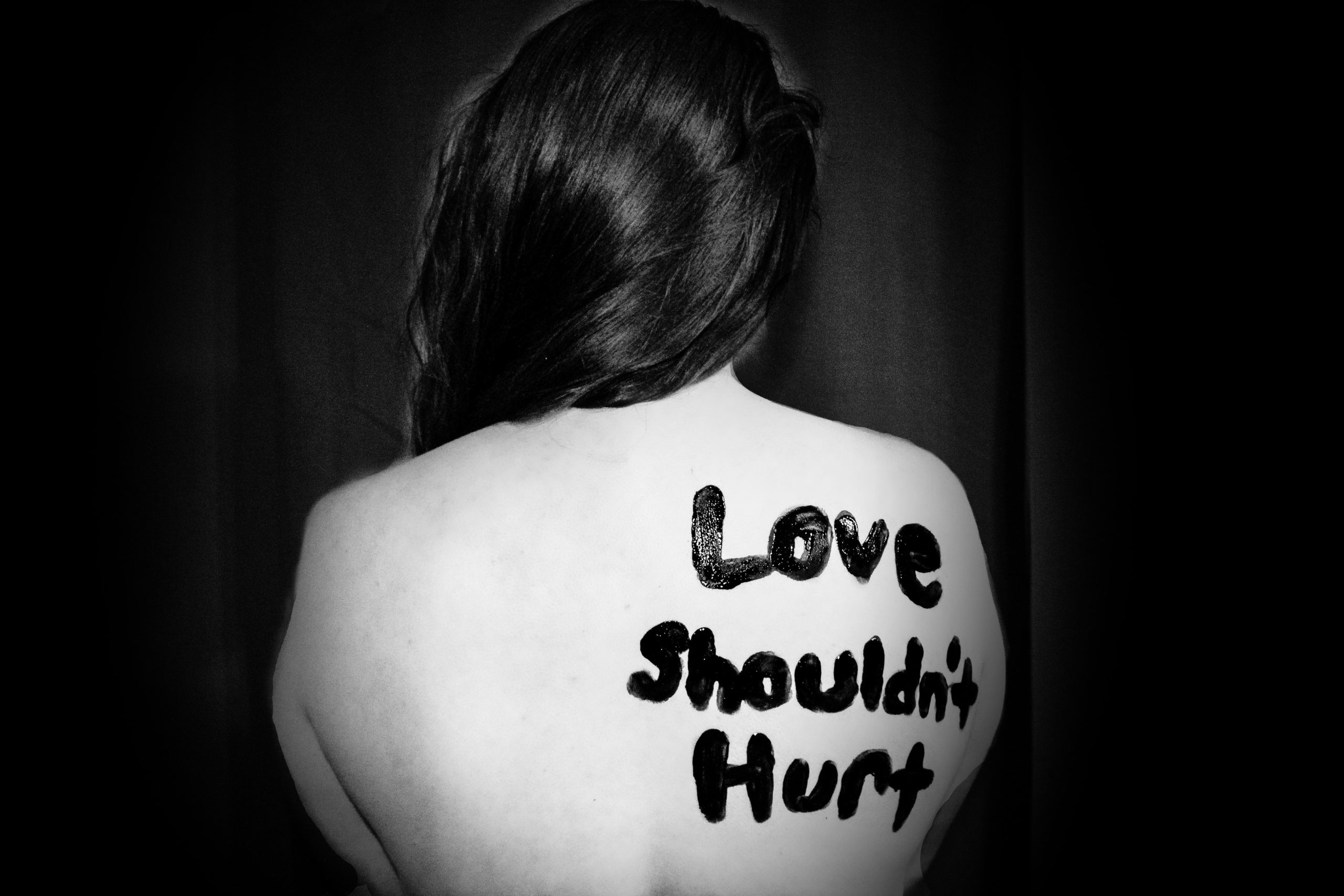 A black and white photo of a woman's back, with "Love shouldn't hurt" painted on her right shoulder blade.