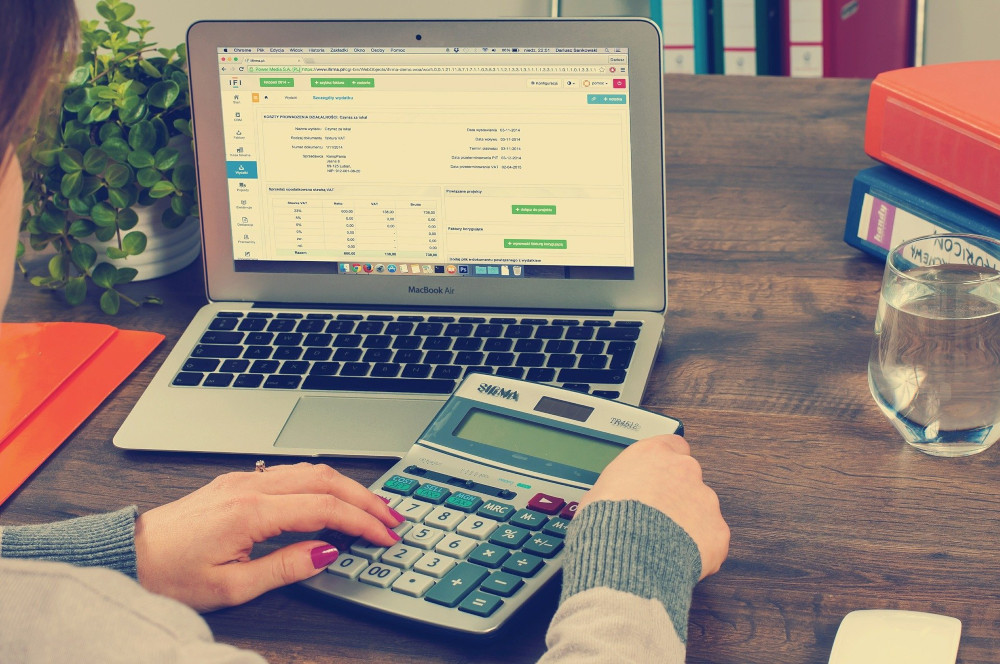 Image captured over shoulder of a person with a manicure who is holding a calculator and looking at the screen of a laptop.