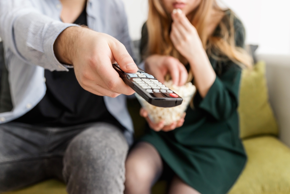 Man points a remote towards the viewer while sitting next to young woman.