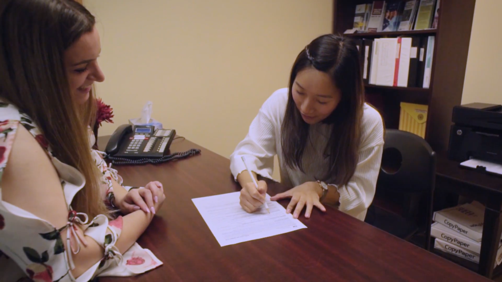 Two women sit at a table while one fills out a form.