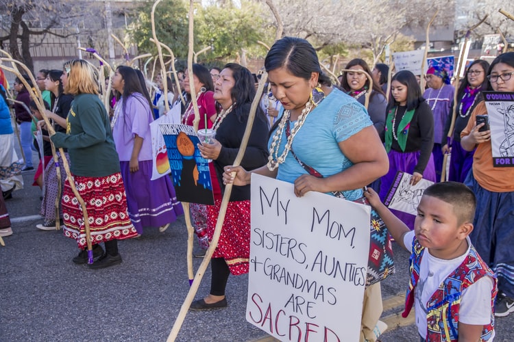 Photo of Protestors, including one woman holding a sign saying "My mom, sisters, aunties + grandmas are sacred."