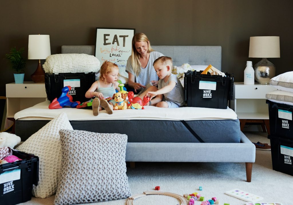 A woman plays with two children on a bed. They are surrounded by moving boxes.